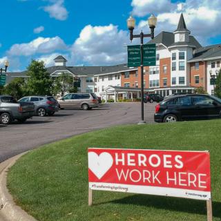 A sign reading 'Heroes Work Here' stands in front of a large building with parked cars in a lot under a partly cloudy sky.