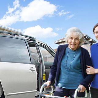 A caregiver assists an elderly woman using a walker near a minivan.