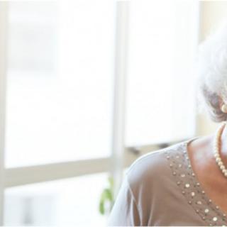 An elderly woman with grey hair and a pearl necklace smiling indoors.