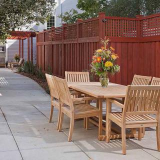 A wooden patio set with chairs and a table, featuring a floral centerpiece, is situated in a garden setting with a red wooden fence.