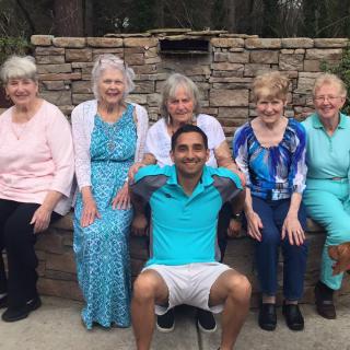 A group of elderly women and a young man sitting and smiling together outdoors.