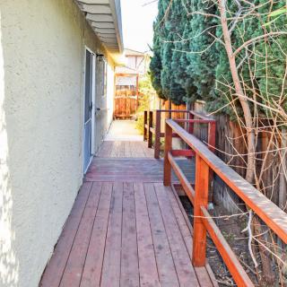 A wooden deck pathway with railings alongside a house, bordered by trees and a fence under sunlight.