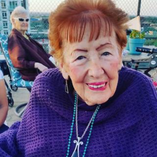 Elderly woman enjoying a day outdoors, wearing a purple shawl and smiling warmly.