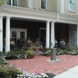 A covered porch with people sitting, surrounded by flowers and a brick walkway.