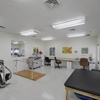 A clean and well-equipped physical therapy room featuring an exercise bike, parallel bars, and a therapist's desk.