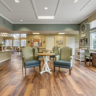 A cozy living room with comfortable chairs and a small table, featuring warm hardwood flooring and bright lighting.
