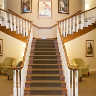 A grand staircase with wooden railings and carpeted steps, flanked by chairs and decorated with artwork.