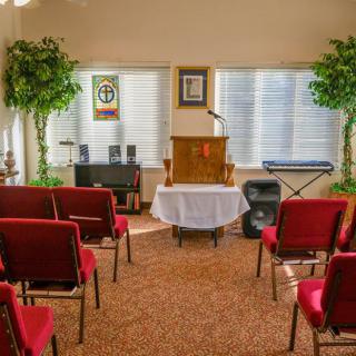 A small, neatly arranged room with red chairs facing a podium and microphone, flanked by potted plants and musical equipment.