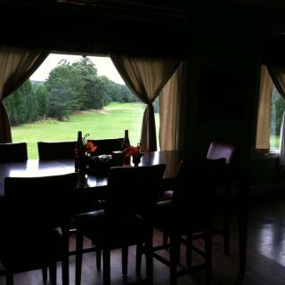 A dimly lit dining room with a large table, chairs, and a view of greenery through the windows.