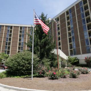 A tall apartment building with an American flag in the foreground, surrounded by well-maintained landscaping and flower beds.