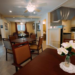 A cozy dining area with wooden tables and chairs, featuring a kitchen in the background and decorative flowers on the table.
