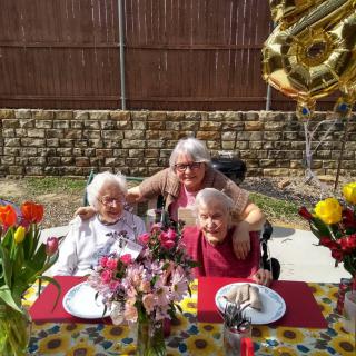Three people celebrating outdoors with balloons and flowers at a decorated table.