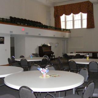 A spacious banquet hall with round tables and chairs, featuring a stage and large windows.