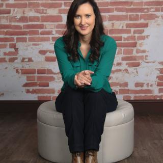 A woman in a green sweater sits on an ottoman against a brick wall backdrop, smiling at the camera.