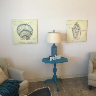 A cozy living room corner with two armchairs, a blue accent table, a lamp, and nautical artwork on the wall.