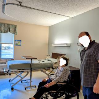 A patient in a wheelchair is seated in a hospital room, with another individual standing nearby.
