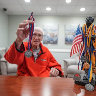 An elderly man proudly displays a medal, with a trophy adorned with multiple medals and an American flag on the table.