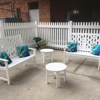 A cozy outdoor patio setup with white benches, small tables, and decorative pillows in a fenced area.