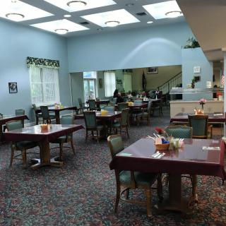 A spacious dining room with multiple tables and chairs, featuring large windows and a carpeted floor.