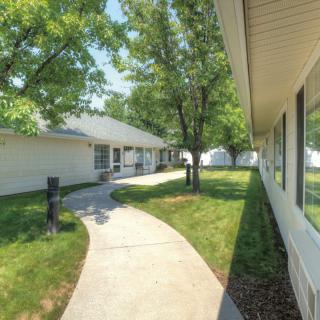 A sunny outdoor view of a residential area with white buildings, a curved sidewalk, green lawns, and trees.