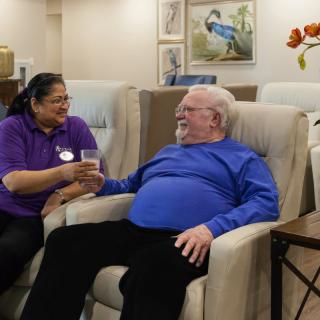 An elderly man sits comfortably in a chair while a caregiver offers him a drink, showcasing a moment of friendly interaction in a care facility.