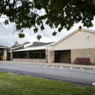 A view of a school building with a parking area and a bench nearby.