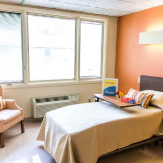 A bright and comfortable hospital room featuring a bed, a chair, and natural light streaming through the windows.