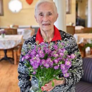 An elderly woman holding a bouquet of purple flowers indoors.