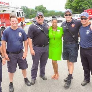 A group of firefighters posing for a photo with a woman in a bright green dress at an outdoor event.