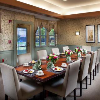 An elegantly decorated dining room with a long table set for a formal meal, featuring cushioned chairs and floral centerpieces.