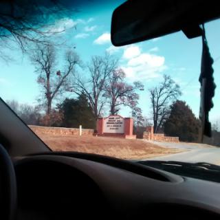 A view from inside a car showing a sign in the distance, with trees and a clear blue sky.