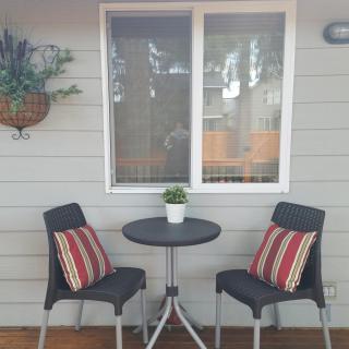 A cozy outdoor patio setup with a small table and two chairs adorned with striped cushions, set against a house wall with a window and hanging plants.
