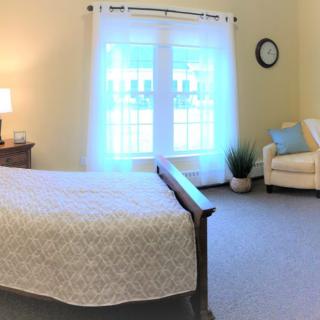A cozy bedroom featuring a bed with a light-colored quilt, an armchair, and large windows with sheer curtains.