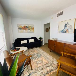 A cozy living room with mid-century modern furnishings, including a black sofa, wooden armchairs, a patterned rug, and decorative artwork on the walls.