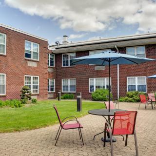A cozy outdoor patio area with tables and chairs, adjacent to a brick building under a partly cloudy sky.
