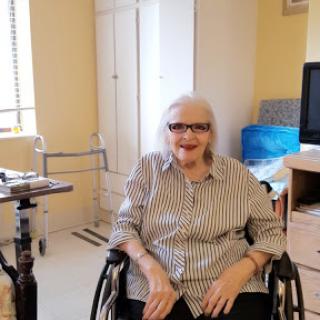 An elderly woman sitting comfortably in a wheelchair, smiling in a cozy indoor setting.