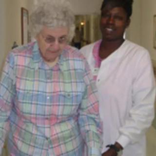 A caregiver assists an elderly woman in walking down a hallway.