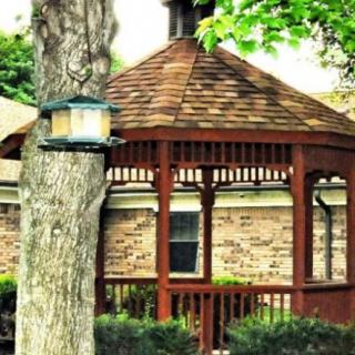 A wooden gazebo situated next to a brick building with a lantern hanging on a nearby tree.