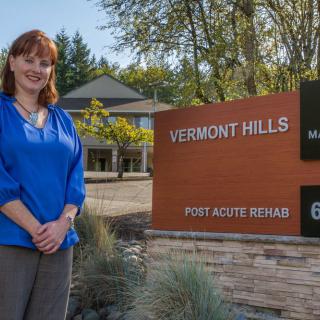 A woman standing next to the Vermont Hills Post Acute Rehab sign on a sunny day.
