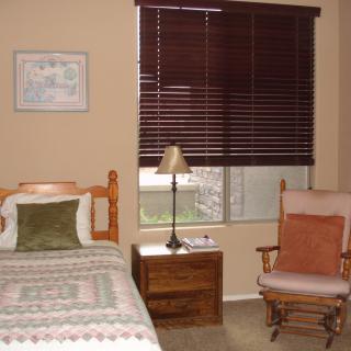 A cozy bedroom featuring a single bed, nightstand with a lamp, window with dark blinds, and a rocking chair.