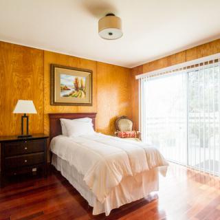 A cozy bedroom with wood paneling and a single bed, illuminated by natural light through a large window.