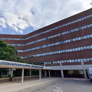 A large hospital building with multiple floors and a canopy at the entrance, set against a partly cloudy sky.