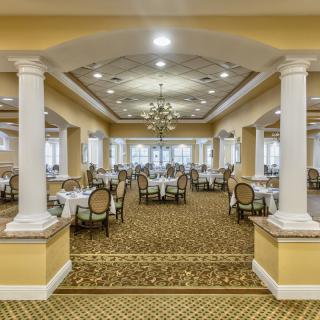 A spacious and elegant dining area featuring chandeliers, pillars, and neatly arranged tables with chairs.