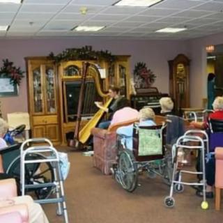 A harpist performs for residents in a nursing home, where elderly individuals sit in wheelchairs and use walkers.