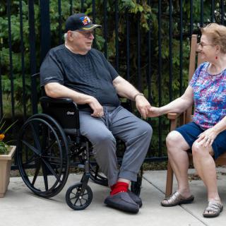 An elderly couple sits outdoors, holding hands. One is in a wheelchair, and they are enjoying each other's company.
