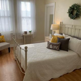 A cozy bedroom featuring a metal bedframe with white bedding, accented by yellow and gray cushions. Natural light filters through the windows, complemented by a bedside lamp. A decorative wreath hangs above the bed.