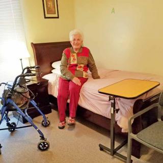 An elderly woman sitting on a bed in a care facility room with a walker nearby.