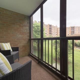 A cozy screened balcony with wicker chairs and outdoor cushions, offering a view of nearby apartment buildings.