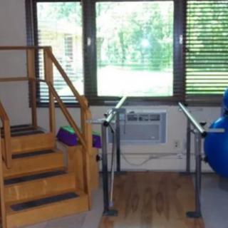 A therapy room equipped with stairs, parallel bars, and exercise ball near large windows.