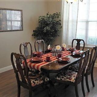 A dining room with a neatly set table, featuring a striped table runner and matching place settings, surrounded by six chairs. A potted plant stands in the corner near a window.